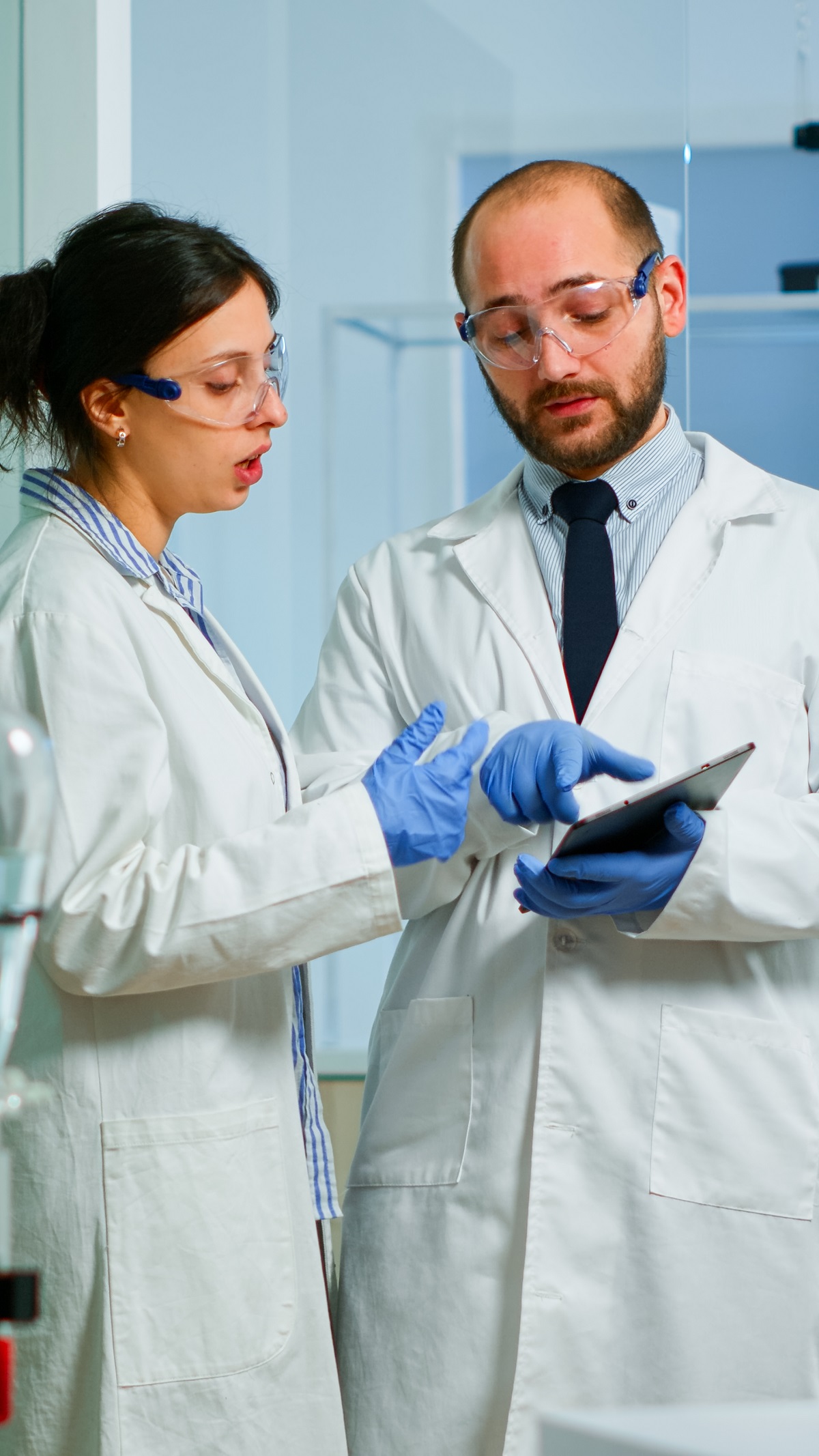 Chemist nurse explainig to doctor vaccine development in modern equipped laboratory pointing on tablet. Team of doctors examining virus evolution using high tech researching diagnosis